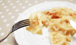 Widok z bliska na widelec z pierogiem z serem, posypanym cynamonem. Na talerzu widać więcej pysznych pierogów.