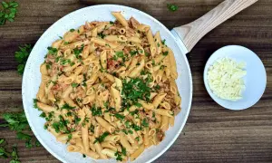 Penne z kremowym sosem i kawałkami pikantnego chorizo, posypane świeżą natką pietruszki. Obok miseczka z tartym parmezanem.