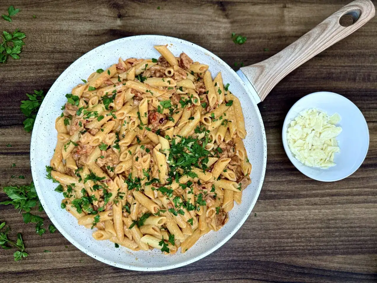 Penne z kremowym sosem i kawałkami pikantnego chorizo, posypane świeżą natką pietruszki. Obok miseczka z tartym parmezanem.